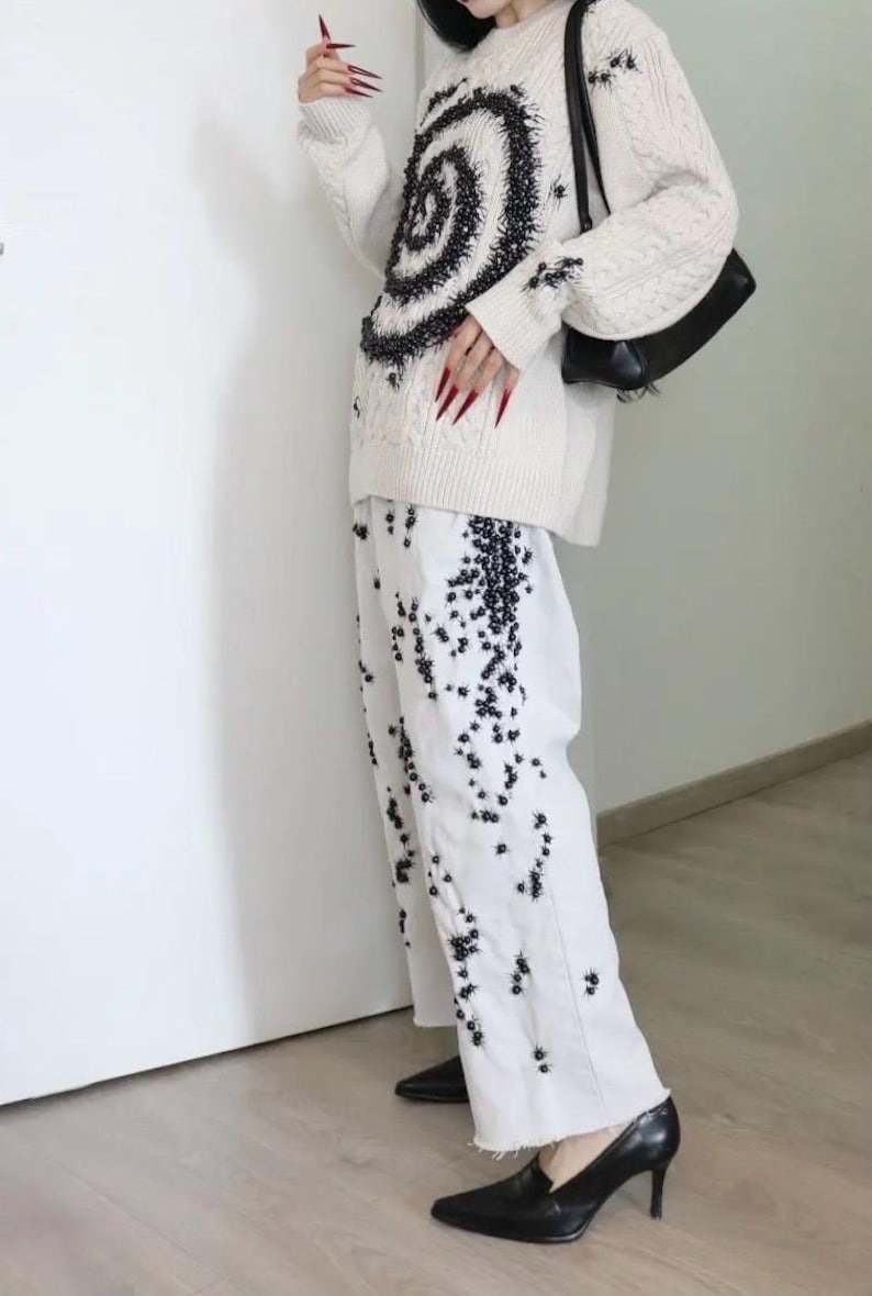 May include: An off-white cable knit sweater featuring a large black spiral pattern and matching black embellishments, paired with white wide-leg trousers decorated with scattered black floral-like embroidery, a black shoulder bag, and black heels.