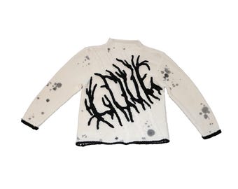 May include: White knitted sweater with a black graphic design of a stylized word. The sweater has a high neck and long sleeves. The sweater has a speckled pattern.
