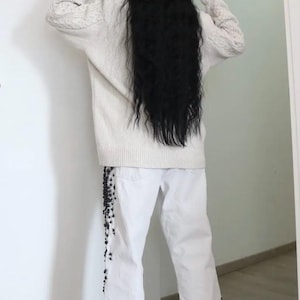 May include: An oversized cream-colored cable knit sweater with long sleeves and white denim jeans featuring black beaded embellishments along the outer leg seams, paired with black pointed-toe heeled shoes.