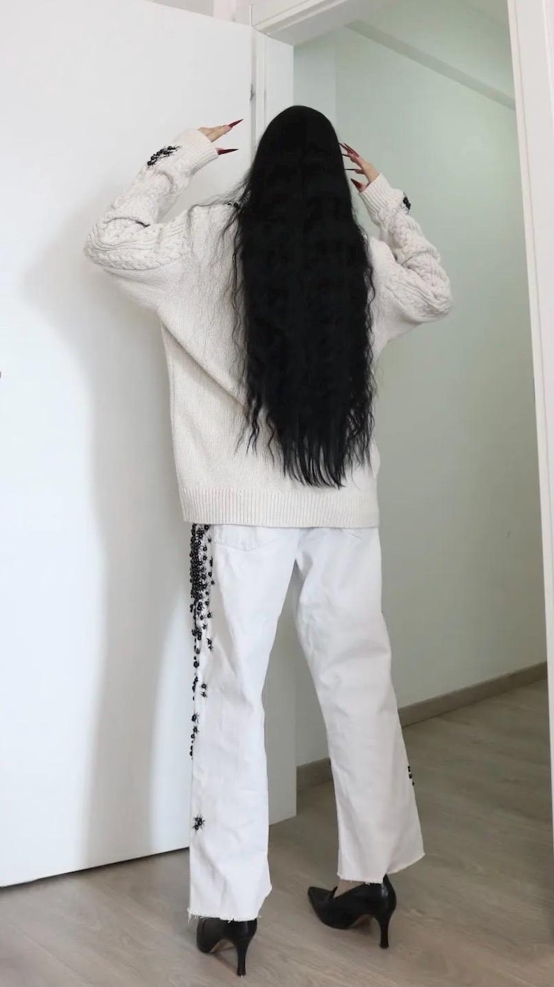 May include: An oversized cream-colored cable knit sweater with long sleeves and white denim jeans featuring black beaded embellishments along the outer leg seams, paired with black pointed-toe heeled shoes.