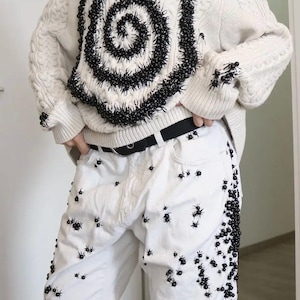 May include: An off-white cable knit sweater featuring a large black spiral design made of small beads, paired with white denim jeans decorated with numerous small black spider-like embellishments and a black belt with a silver buckle.