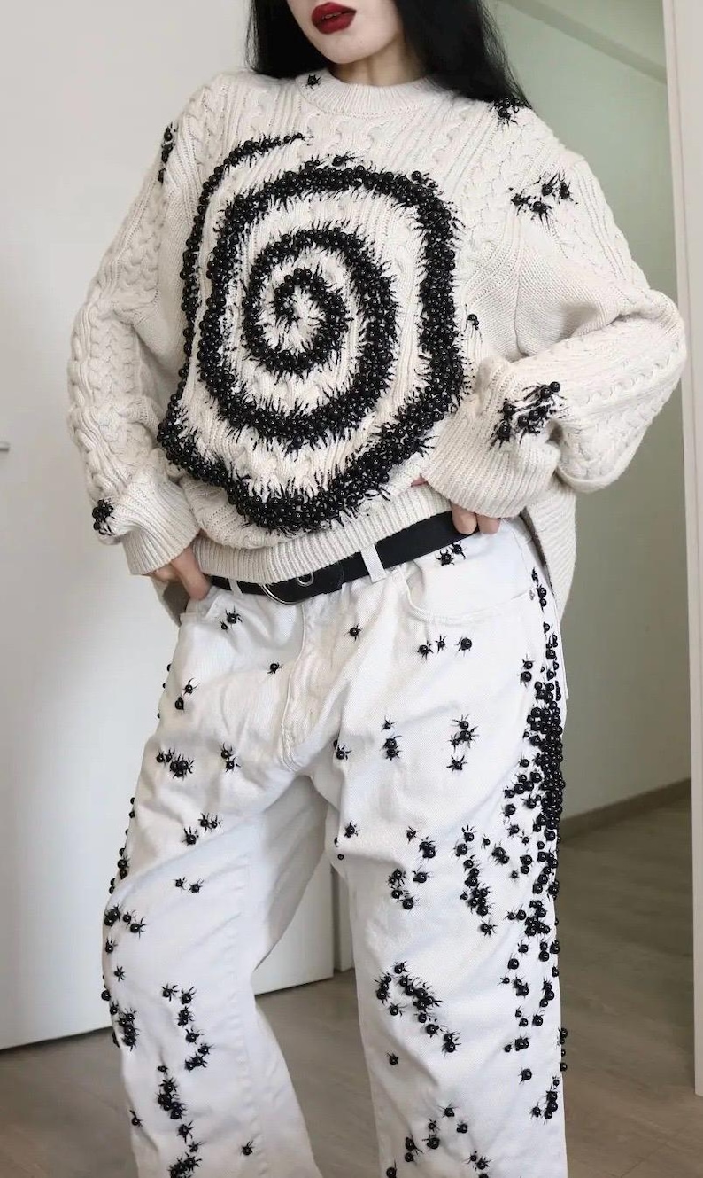 May include: An off-white cable knit sweater featuring a large black spiral design made of small beads, paired with white denim jeans decorated with numerous small black spider-like embellishments and a black belt with a silver buckle.