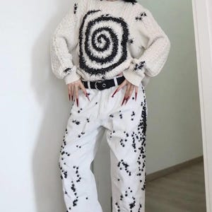 May include: An off-white cable knit sweater featuring a large black spiral design on the front, paired with white denim jeans decorated with numerous small black spider-like embellishments and a black belt with a silver buckle.