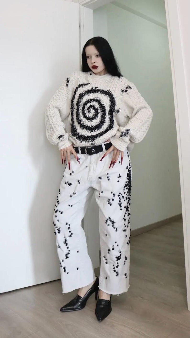 May include: An off-white cable knit sweater featuring a large black spiral design on the front, paired with white denim jeans decorated with numerous small black spider-like embellishments and a black belt with a silver buckle.