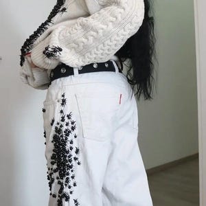 May include: A cream-colored cable knit sweater featuring black beaded spider embellishments along the front edge and sleeves, paired with white denim jeans decorated with clusters of black beaded spiders and a black belt with silver grommets.