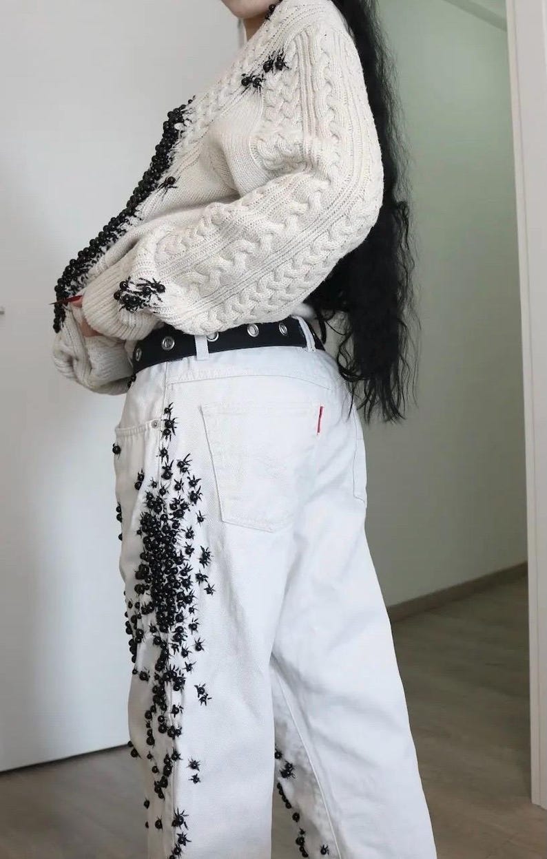 May include: A cream-colored cable knit sweater featuring black beaded spider embellishments along the front edge and sleeves, paired with white denim jeans decorated with clusters of black beaded spiders and a black belt with silver grommets.