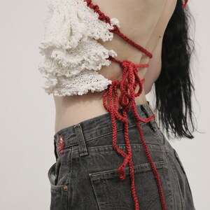 May include: A white crochet top with red crocheted straps tied at the back. The top has a ruffled design and is worn with dark denim jeans.