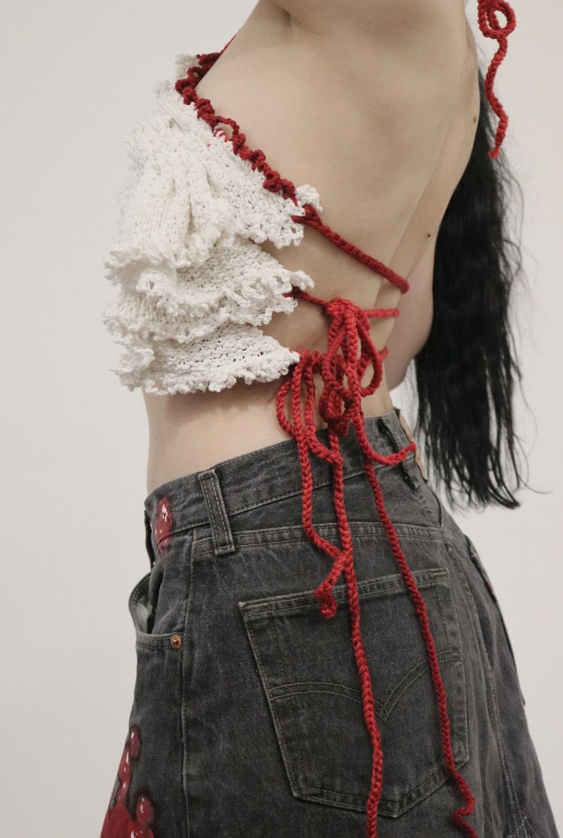 May include: A white crochet top with red crocheted straps tied at the back. The top has a ruffled design and is worn with dark denim jeans.