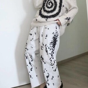May include: A cream-colored cable knit sweater featuring a large black spiral design and scattered black embellishments, paired with white wide-leg trousers decorated with a dense pattern of small black beaded spider-like motifs and black pointed shoes.