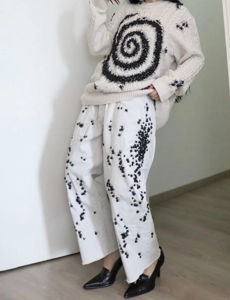 May include: A cream-colored cable knit sweater featuring a large black spiral design and scattered black embellishments, paired with white wide-leg trousers decorated with a dense pattern of small black beaded spider-like motifs and black pointed shoes.