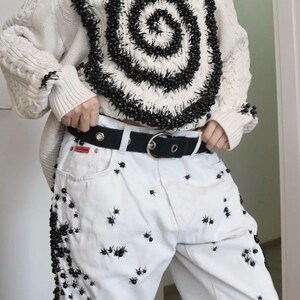 May include: A cream-colored cable knit sweater featuring a large black spiral design made of small beads, paired with white denim jeans decorated with scattered black bead embellishments and a black belt with a silver-toned metal buckle.