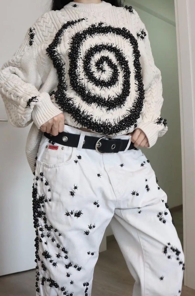 May include: A cream-colored cable knit sweater featuring a large black spiral design made of small beads, paired with white denim jeans decorated with scattered black bead embellishments and a black belt with a silver-toned metal buckle.
