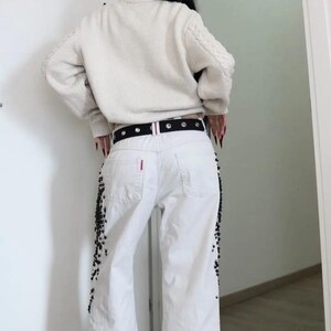 May include: A cream-colored cable knit sweater paired with white denim jeans featuring black beaded embellishments along the outer seams. The jeans are worn with a black belt featuring silver-toned grommets and a small red tag on the back pocket.