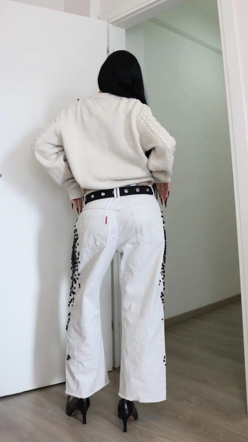 May include: A cream-colored cable knit sweater paired with white denim jeans featuring black beaded embellishments along the outer seams. The jeans are worn with a black belt featuring silver-toned grommets and a small red tag on the back pocket.