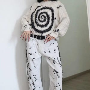 May include: A cream-colored cable knit sweater featuring a large black spiral design and scattered black spider motifs, paired with white denim jeans decorated with numerous small black spider embellishments and a black belt with a silver buckle.