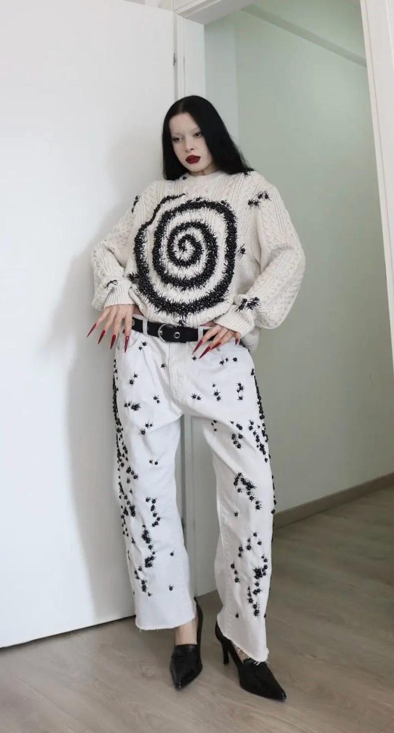 May include: A cream-colored cable knit sweater featuring a large black spiral design and scattered black spider motifs, paired with white denim jeans decorated with numerous small black spider embellishments and a black belt with a silver buckle.