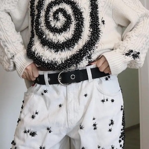 May include: An off-white cable knit sweater featuring a large black spiral design made of small beads, paired with white denim jeans. Both the sweater and the jeans are embellished with numerous small black spider-like bead ornaments. A black belt with a silver buckle is worn at the waist.