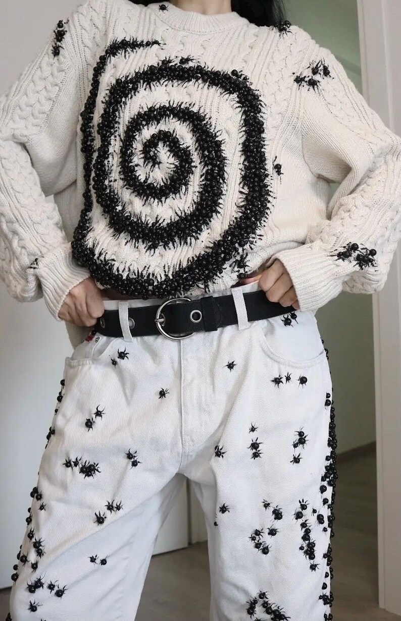 May include: An off-white cable knit sweater featuring a large black spiral design made of small beads, paired with white denim jeans. Both the sweater and the jeans are embellished with numerous small black spider-like bead ornaments. A black belt with a silver buckle is worn at the waist.