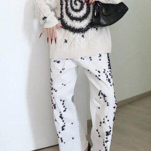 May include: A cream-colored cable knit sweater featuring a large black spiral design and scattered black embellishments, paired with white wide-leg denim jeans decorated with numerous small black floral-like accents and a black shoulder bag.
