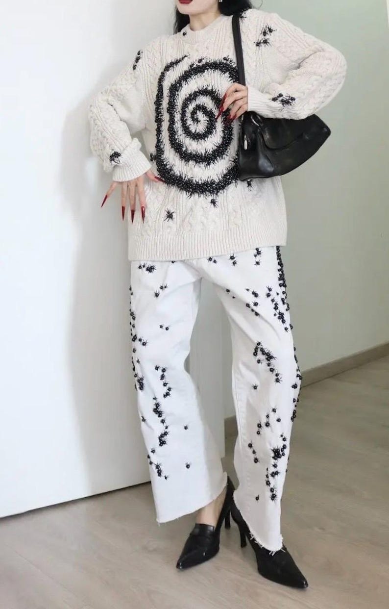 May include: A cream-colored cable knit sweater featuring a large black spiral design and scattered black embellishments, paired with white wide-leg denim jeans decorated with numerous small black floral-like accents and a black shoulder bag.
