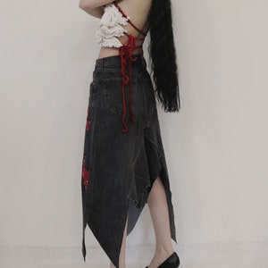 May include: A woman wearing a white crocheted top with red straps and a black denim skirt with red accents. The skirt is asymmetrical and has a long, flowing train.