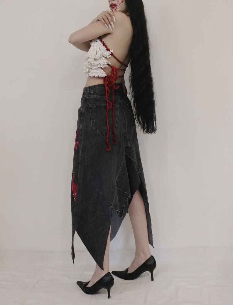 May include: A woman wearing a white crocheted top with red straps and a black denim skirt with red accents. The skirt is asymmetrical and has a long, flowing train.