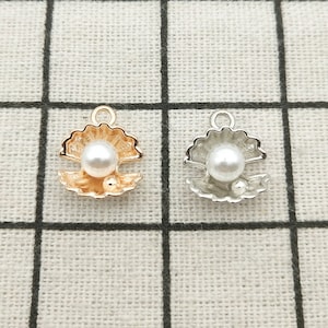 May include: Two gold and silver clam shell charms with a white pearl inside. The charms have a loop at the top for attaching to jewelry.
