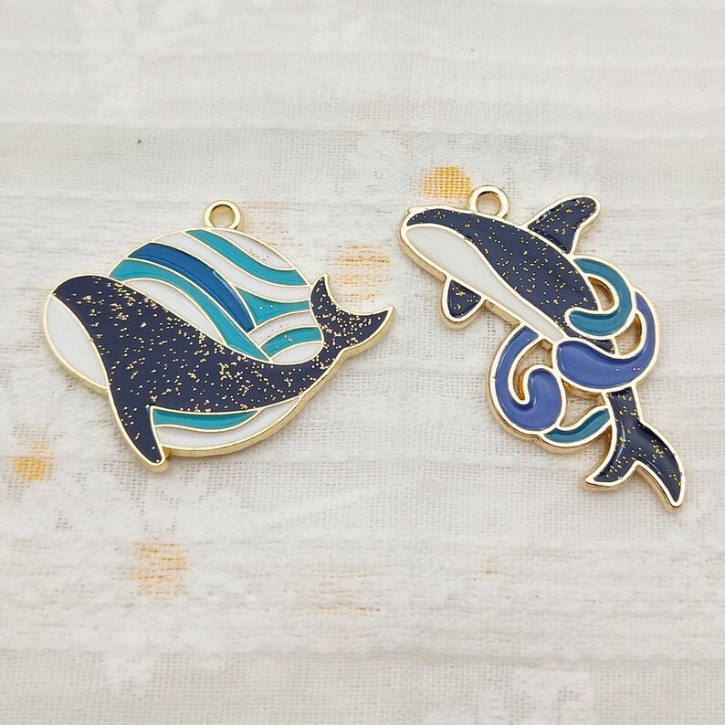 May include: Two gold-tone metal charms featuring a whale design. One charm is a whale with blue and white waves, the other is a whale with a blue and white swirl design.