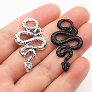 Puede incluir: Dos dijes con forma de serpiente, uno con acabado plateado metálico y otro con acabado negro mate. Cada dije presenta un cuerpo enroscado con un patrón texturizado similar a escamas en la superficie y una cabeza triangular distintiva.