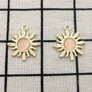 Puede incluir: Dos colgantes de sol de color dorado con un centro rosa. Los colgantes tienen un dise&ntilde;o detallado con rayos que emanan del centro.