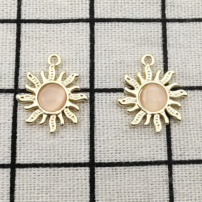 Puede incluir: Dos colgantes de sol de color dorado con un centro rosa. Los colgantes tienen un dise&ntilde;o detallado con rayos que emanan del centro.