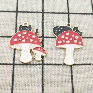 May include: Two gold-tone metal charms shaped like mushrooms with red tops and white spots. One charm features a black cat sitting on top of the mushroom, and the other charm features a black cat sleeping on top of the mushroom.