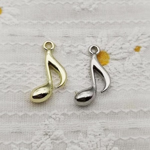 Op de afbeelding: Twee muzieknootbedels, één met een gepolijste goudkleurige afwerking en de andere met een gepolijste zilverkleurige afwerking. Elke bedel heeft een kleine ronde lus aan de bovenkant voor bevestiging, met een glad, gebogen en reflecterend oppervlak.