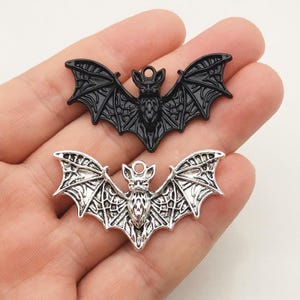 May include: Two bat-shaped charms, one in a solid matte black finish and one in a metallic silver finish. Each charm features detailed wing structures with intricate webbing patterns and a small circular loop at the top for attachment.