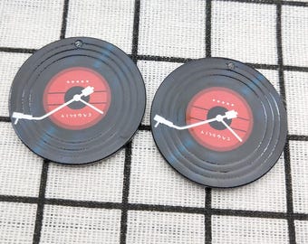 May include: Two black vinyl record-shaped charms with a red centre and white stylus. The centre of each charm has white text and five white dots. The charms have a small hole at the top for hanging.