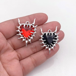 May include: Two heart-shaped pendants featuring a faceted red or black gemstone set within a silver-toned metal frame. The frame is designed with sharp, thorn-like protrusions extending outward and two small horn-like points at the top.