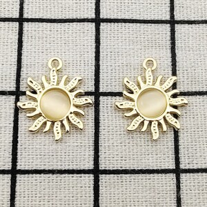 Puede incluir: Dos colgantes de sol de tono dorado con un centro blanco opalescente. Los colgantes tienen un dise&ntilde;o detallado con rayos que emanan del centro.