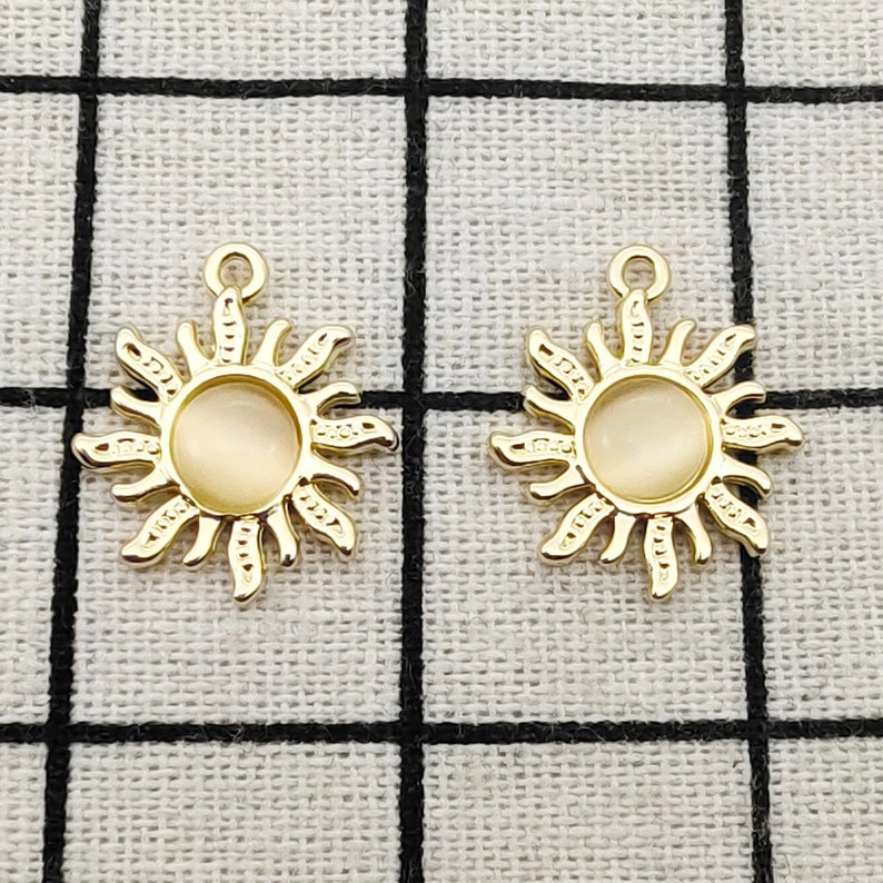 Puede incluir: Dos colgantes de sol de tono dorado con un centro blanco opalescente. Los colgantes tienen un dise&ntilde;o detallado con rayos que emanan del centro.