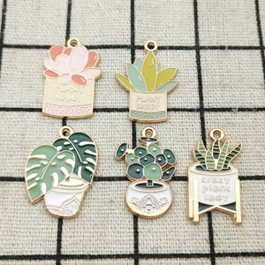 May include: Five gold-colored metal charms shaped like potted plants. The charms feature different plant designs, including a succulent, a monstera, and a cactus. Each charm has a different text label, including "Plant Mama", "Plant Daddy", "Crazy Plant Lady".