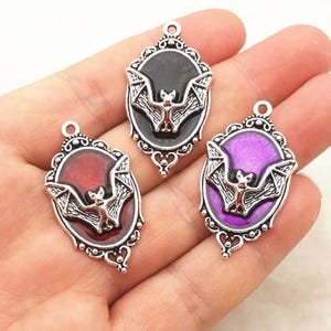 May include: Three oval-shaped metal pendants featuring a silver-toned bat relief centered on a colored background. Each pendant has an ornate, filigree-style frame with a top loop. The backgrounds are solid black, deep red, and vibrant purple.
