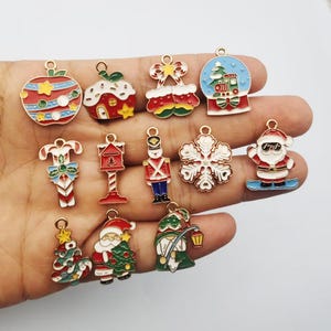May include: A collection of twelve metal enamel charms featuring Christmas themes including a striped ornament, a decorated house, stockings, a snow globe with a train, a candy cane, a mailbox, a nutcracker, a snowflake, Santa Claus, and a gnome.