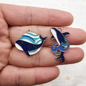 May include: Two gold-tone metal charms featuring blue and white enamel whale designs. One charm depicts a whale swimming in a circle of blue and white waves. The other charm depicts a whale jumping out of the water with a blue and white wave behind it.