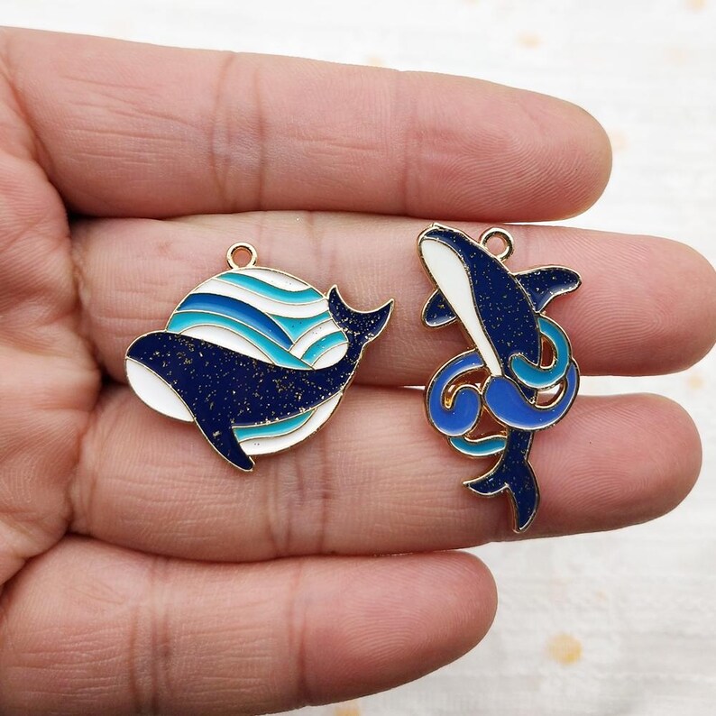 May include: Two gold-tone metal charms featuring blue and white enamel whale designs. One charm depicts a whale swimming in a circle of blue and white waves. The other charm depicts a whale jumping out of the water with a blue and white wave behind it.