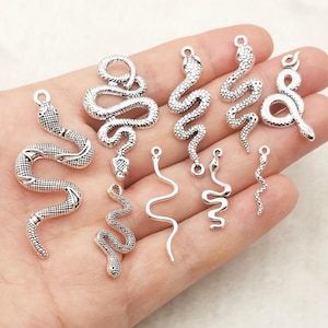 Puede incluir: Una colección de dijes de serpiente de metal en tono plateado de varios tamaños y diseños. Cada dije cuenta con una anilla en la parte superior para su fijación, algunos con texturas de escamas detalladas y otros con una silueta serpenteante lisa.