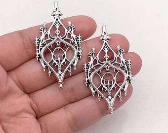 May include: Two silver-toned metal pendants featuring an intricate, openwork design with pointed, cathedral-like architectural elements and a central diamond-shaped cutout. The metal has a polished finish with dark recessed areas to highlight the details.