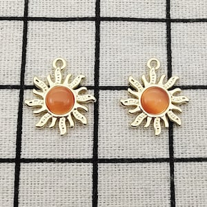Puede incluir: Dos colgantes de sol de color dorado con centros de vidrio naranja. Los colgantes est&aacute;n sobre un fondo de tela blanca.