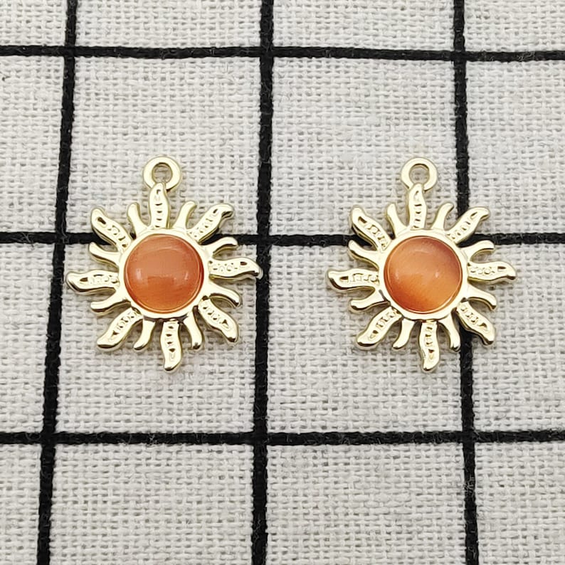 Puede incluir: Dos colgantes de sol de color dorado con centros de vidrio naranja. Los colgantes est&aacute;n sobre un fondo de tela blanca.