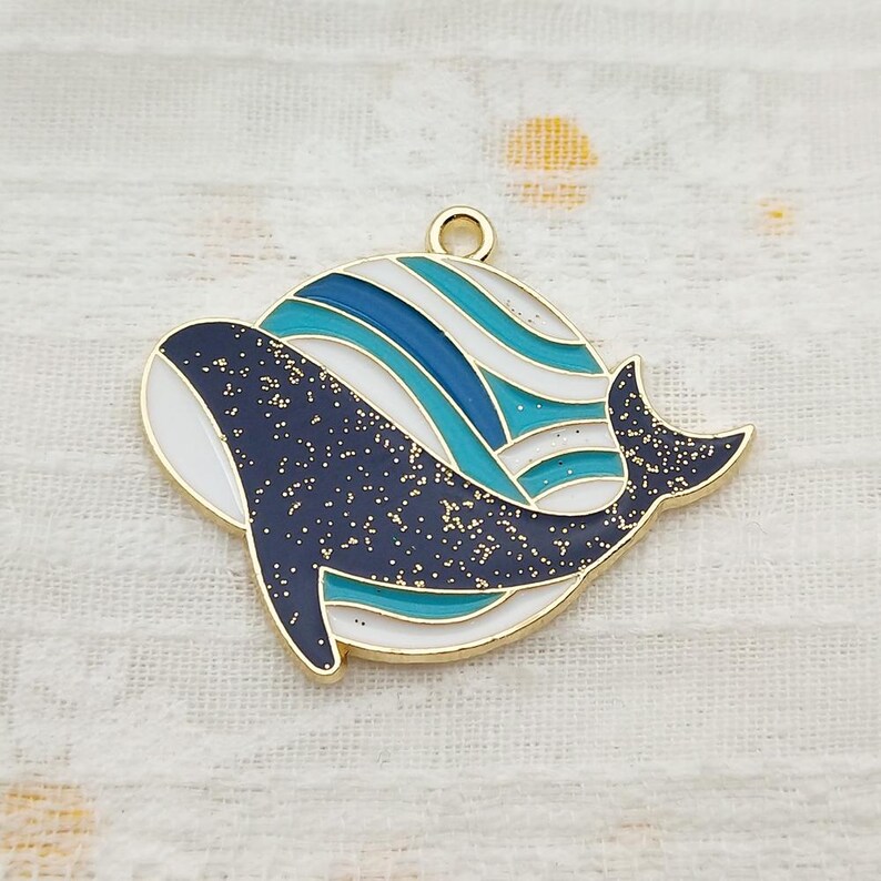 May include: Gold-tone charm featuring a blue and white striped circle with a black whale with gold glitter inside. The whale is swimming through the circle.
