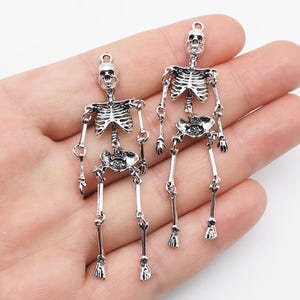 Peut inclure: Deux breloques squelette en métal argenté avec des articulations mobiles au niveau des épaules, des hanches et des genoux. Chaque squelette possède une petite boucle circulaire au sommet du crâne pour la fixation, avec une cage thoracique détaillée.