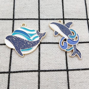 May include: Two gold-tone metal whale charms. One charm features a blue and white whale with waves, the other charm features a blue whale with a blue tail fin.